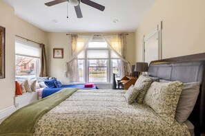 A bedroom with a large bed, a desk, a chair, a ceiling fan, and a window with curtains.