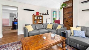 Ein Wohnzimmer mit einem grauen Ecksofa, einem Holztisch, einem Bücherregal und einer Wand mit Ziegeln.