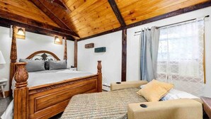 Ein Schlafzimmer mit Holzbalkendecke, einem Bett mit Holz Kopfteil, einem Nachttisch, einem Sessel und einem Fenster mit Vorhängen.