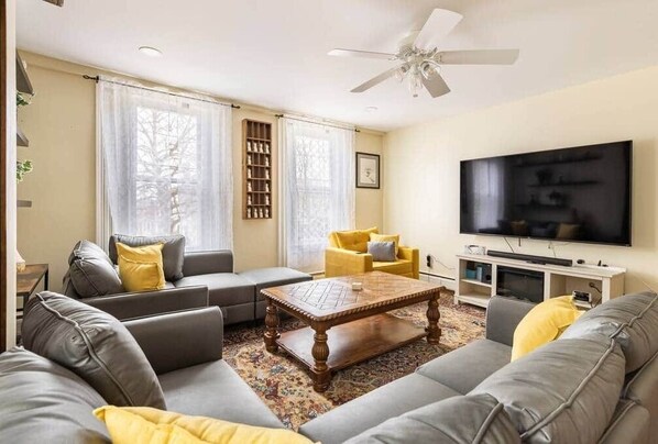 Una sala de estar con un sofá gris, una mesa de centro de madera, un sillón amarillo y un televisor de pantalla plana.
