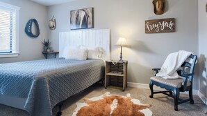 Un dormitorio con una cama, una silla, una mesita de noche, una lámpara, un tapete de cuero de vaca y adornos en la pared, incluyendo un letrero con la frase 'hey yall' y un sombrero de vaquero.