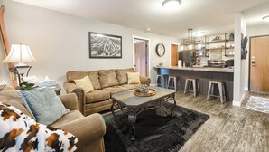 Una sala de estar con sofá, mesa de centro y una zona de cocina con taburetes y barra.