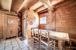 A wooden cabin interior with a dining table, chairs, a cabinet, a window, and a red bucket labeled 'INCENDI'. - Chalet “6p Classique - Clématite” with Mountain View, Private Terrace & Wi-Fi (Verchaix)