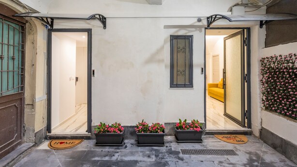 Entrada de hotel con tres plantas en macetas, un felpudo y un letrero en la pared.