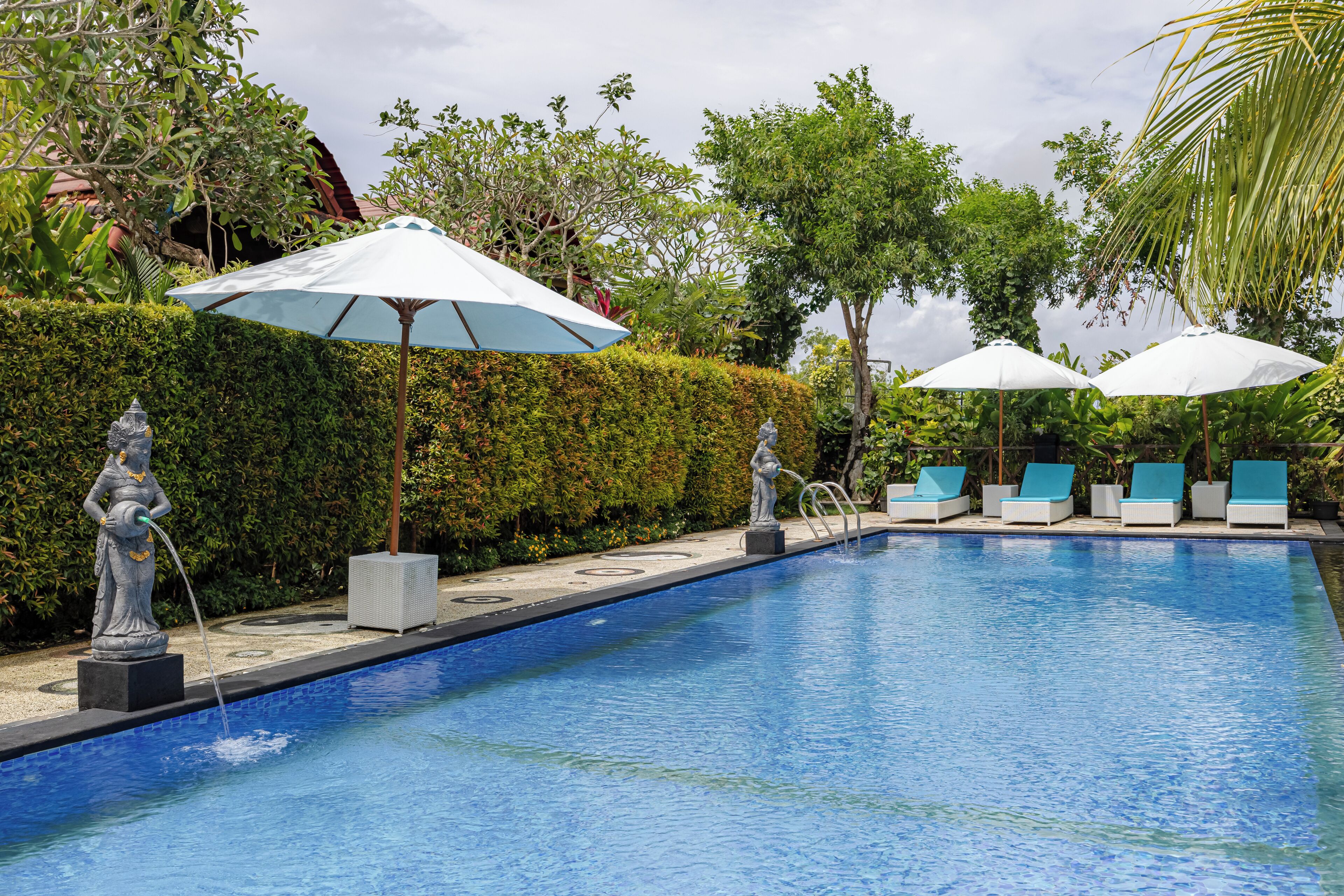 A swimming pool with lounge chairs and umbrellas, surrounded by greenery and statues.