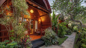 Une maison en bois dotée d’un porche, entourée d’une végétation luxuriante et bordée d’un chemin de gravier aux motifs circulaires.