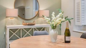 A round wooden table with a white vase and a bottle of wine, a mirror, and two lamps on a white console table. - Gatehouse Cottage, Cartmel (Cartmel)