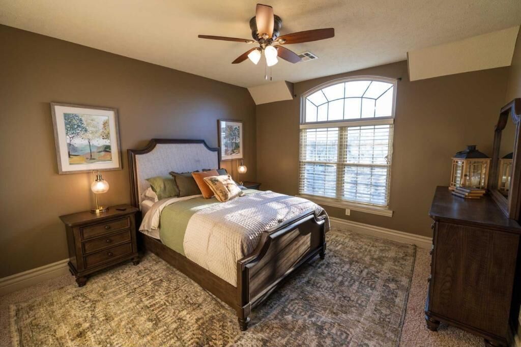 A bedroom with a large bed, a ceiling fan, a window with blinds, and a nightstand with a lamp.