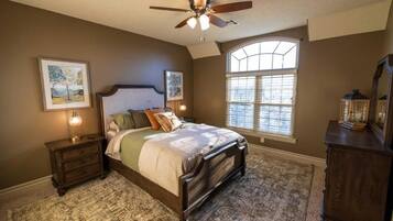 A bedroom with a large bed, a ceiling fan, a window with blinds, and a nightstand with a lamp.
