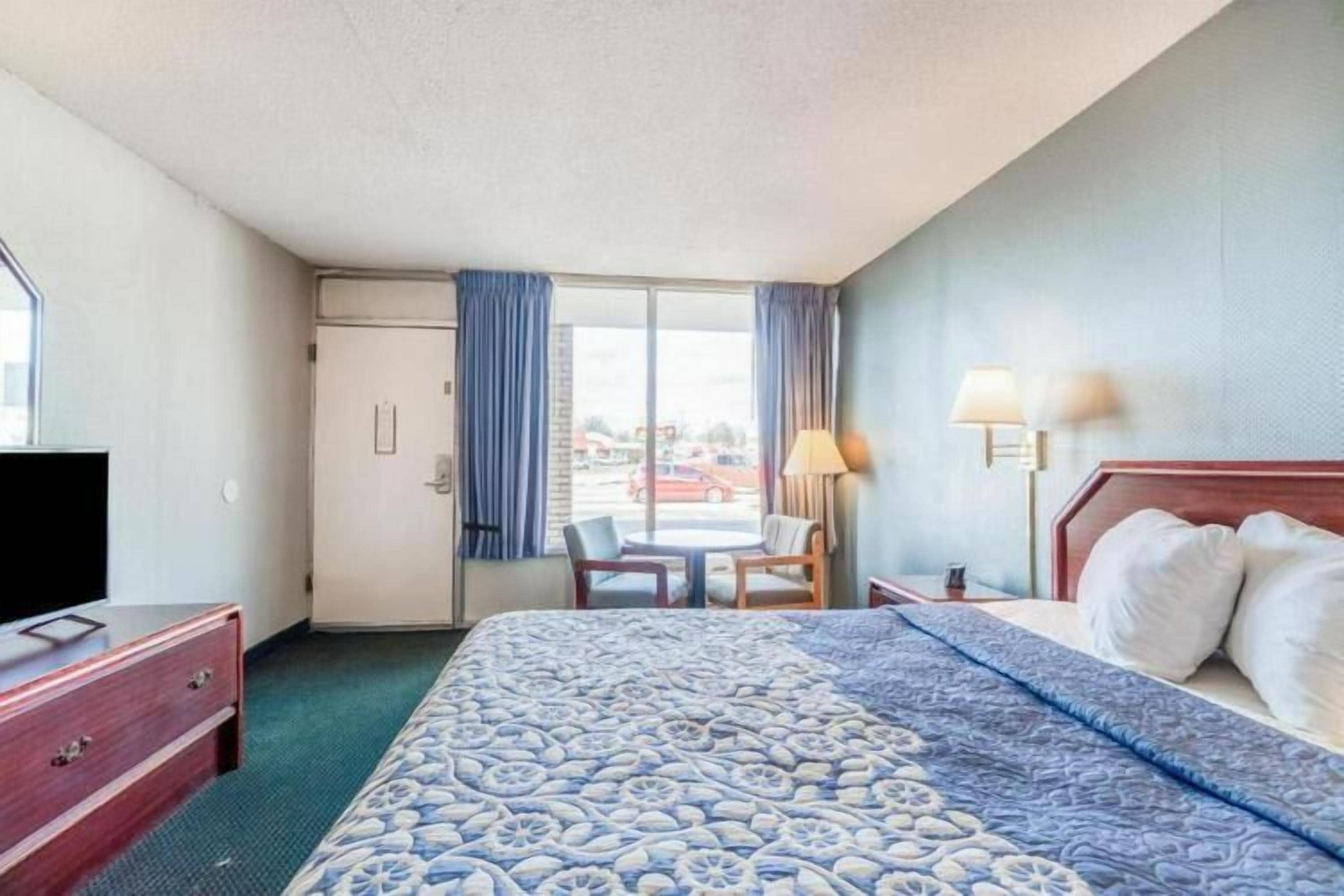 Una habitación de hotel con cama, escritorio, silla, televisor y ventana con cortinas.