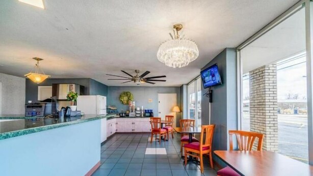 Un hall d’hôtel comprenant un comptoir d’accueil, un espace repas avec des chaises et une table, un lustre, des ventilateurs de plafond et un téléviseur fixé au mur.