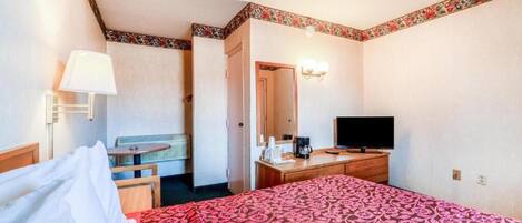Quarto de hotel com cama, escrivaninha, cadeira, televisão e luminária.