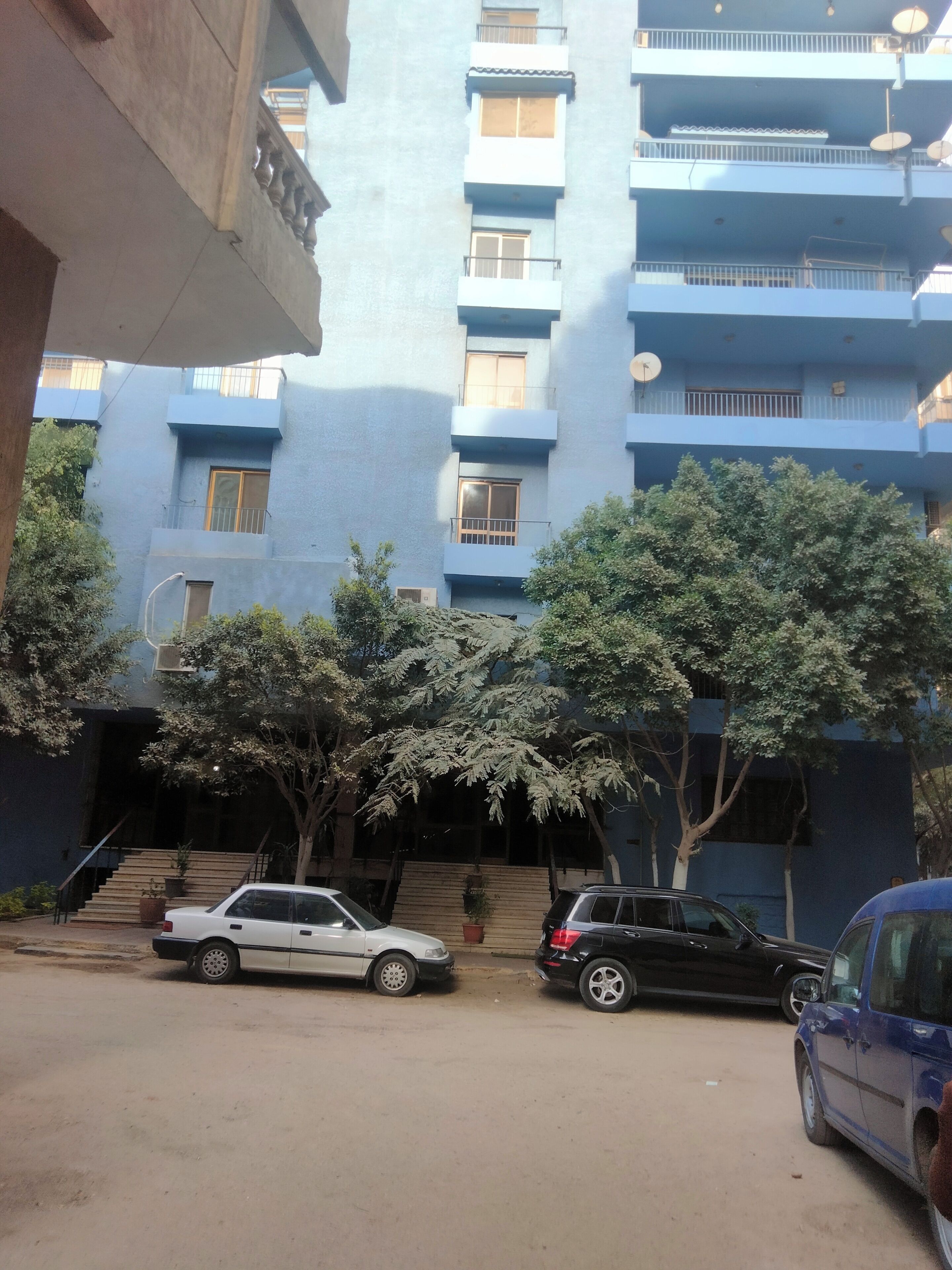 Un edificio de varios pisos con balcones, un automóvil estacionado al frente y árboles en primer plano.