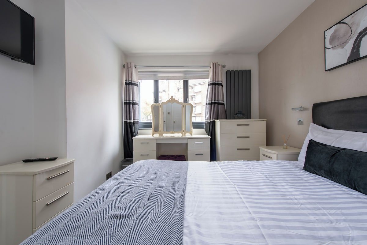 A bedroom with a bed, a dresser, a vanity with a mirror, a TV mounted on the wall, and a window with curtains.