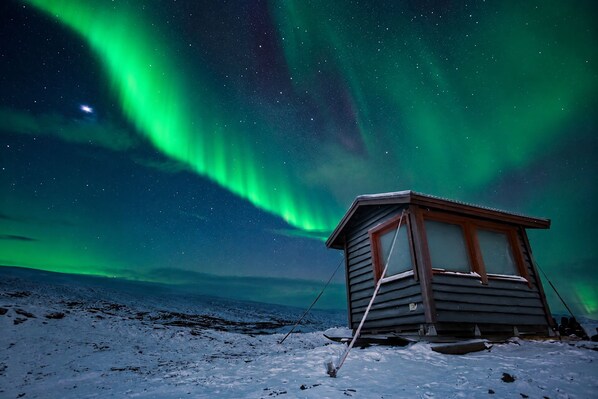 A wooden cabin with a window and a door, situated on a snow-covered ground with a starry night sky and aurora borealis in the background.