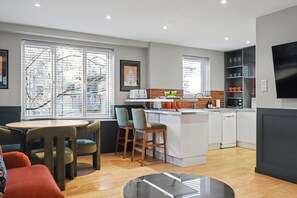 Una cocina moderna con armarios blancos, microondas integrado y una pequeña isla. Hay dos taburetes de bar y una mesa redonda con sillas.