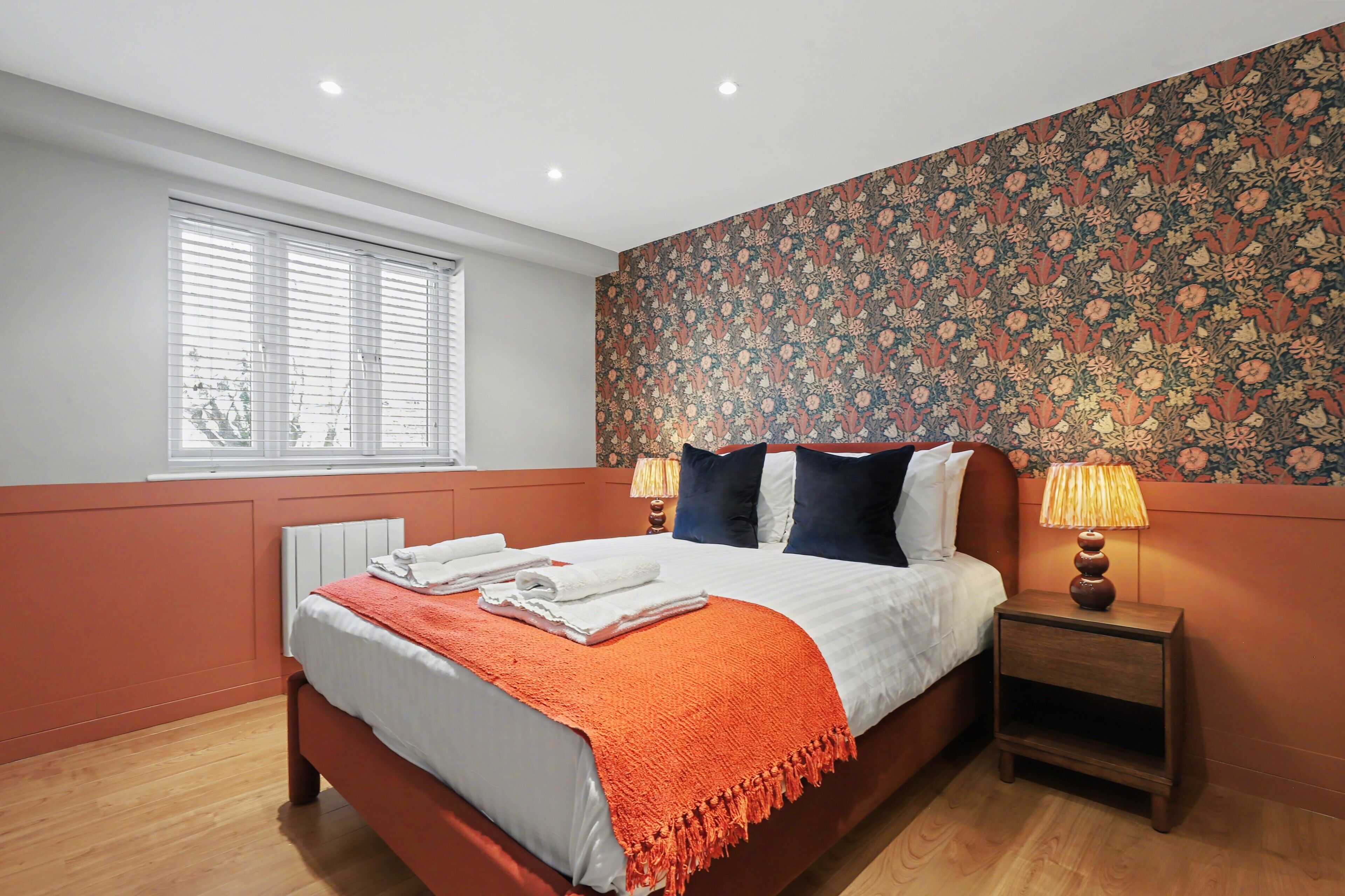 Quarto com cama, duas mesas de cabeceira com luminárias, janela com persianas e papel de parede floral.