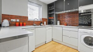 Une cuisine moderne avec des meubles blancs, un lave-vaisselle, un four à micro-ondes et un lave-linge. Il y a une fenêtre avec des stores et un pare-feu en briques.