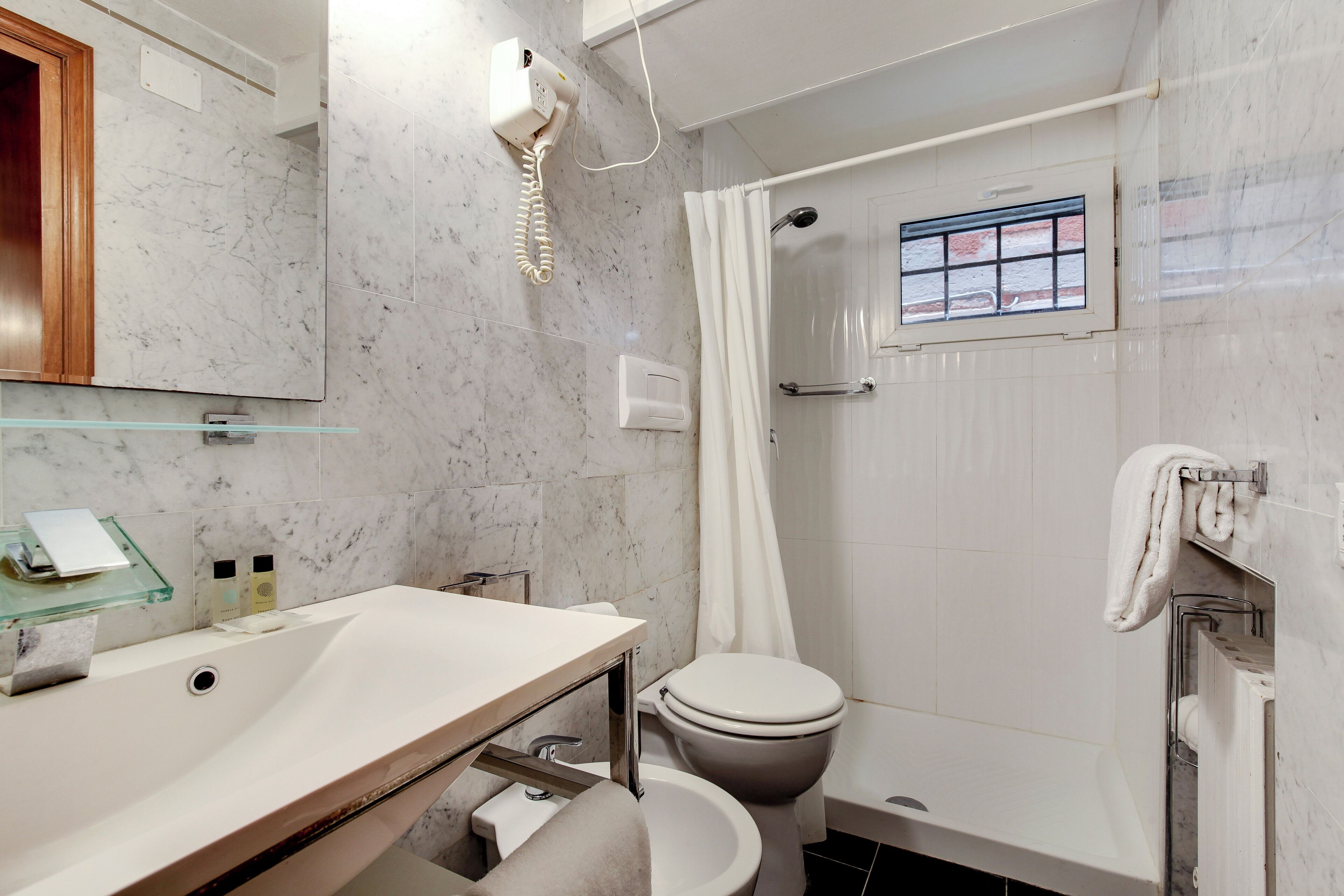 Een badkamer met een marmeren wastafel, een toilet, een douche met raam en een handdoekrek.