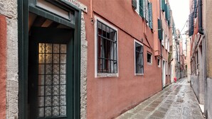 A narrow cobblestone alley with buildings on either side, featuring a door with a grid pattern and a window with a metal grille. - Leo Apartment (Venezia)