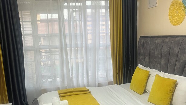 Una cama bien hecha con ropa de cama blanca y amarilla, un cabecero gris y vistas al exterior a través de cortinas de encaje.