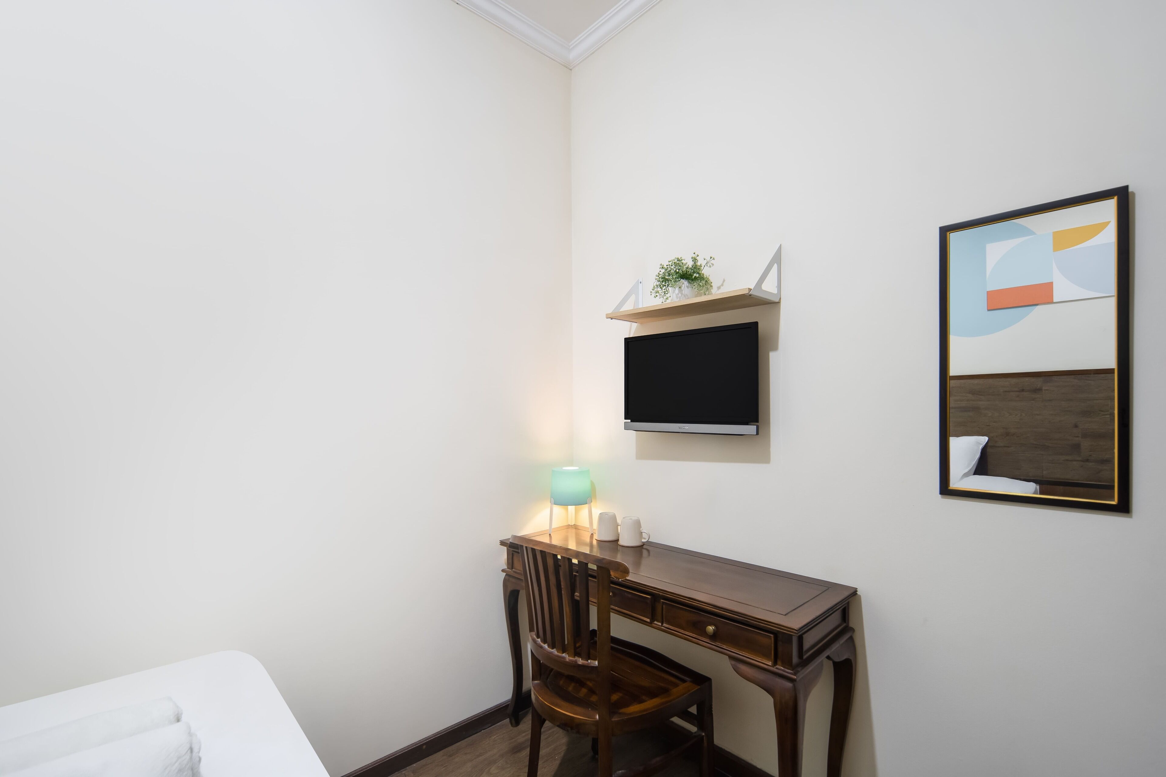 A hotel room with a bed, a desk with a chair, a TV, a shelf with a plant, and a framed picture on the wall.