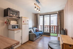 A compact kitchen with a microwave, toaster, and a small refrigerator. There is a wooden dining table and a sofa with a view to a balcony. - Le Petit Chez Nous: Ideal City Break with Balcony, Comfort & Tranquility (Villard-de-Lans)