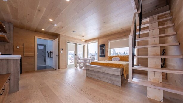 Une pièce spacieuse avec un plancher en bois, un lit recouvert d’une courtepointe jaune, un coin repas avec une table et des chaises, et un escalier avec des marches en bois.