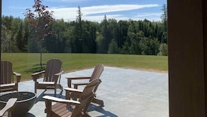 Une terrasse extérieure avec des fauteuils Adirondack et un foyer, donnant sur une pelouse et une forêt.