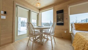 L’intérieur chaleureux d’un chalet avec une table et des chaises en bois, un lit recouvert d’une couverture jaune et de grandes fenêtres donnant sur un paysage enneigé.