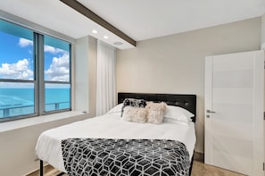 A modern bedroom with a large window, a bed with patterned bedding, and a view of the ocean. - Beautiful Ocean front Condo Hollywood (Hollywood)