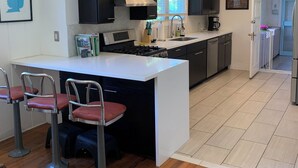 Une cuisine moderne avec des meubles de couleur sombre, un îlot central blanc doté de tabourets, une cuisinière encastrée et une fenêtre avec des stores.