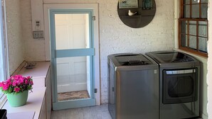 Une buanderie équipée d’un lave-linge et d’un sèche-linge, une porte avec un cadre bleu, une étagère fixée au mur avec des bocaux, et des guirlandes lumineuses au plafond.
