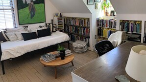 Une chambre mansardée chaleureuse avec un lit en mezzanine, une petite table ronde avec des livres, une bibliothèque et une grande fenêtre avec des stores.