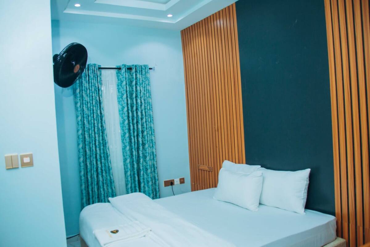 Habitación de hotel con cama, cortinas, ventilador de pared y panel de pared de madera.