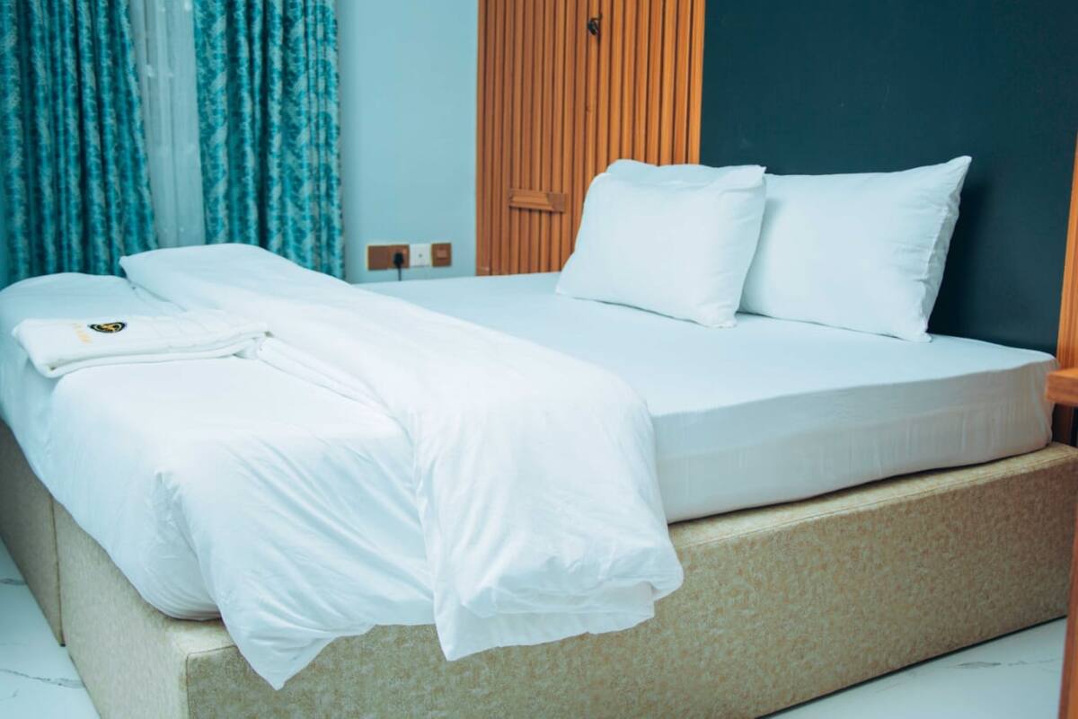 Una cama bien hecha con sábanas y almohadas blancas, un cabecero de madera y una ventana con cortinas azules.
