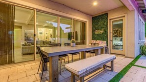 Uma área de refeições ao ar livre com uma mesa comprida, bancos e cadeiras, anexa a uma porta de vidro deslizante que dá para um interior moderno.