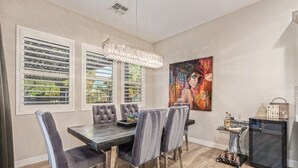Uma sala de jantar com uma mesa, cadeiras, um lustre, um quadro e uma geladeira.