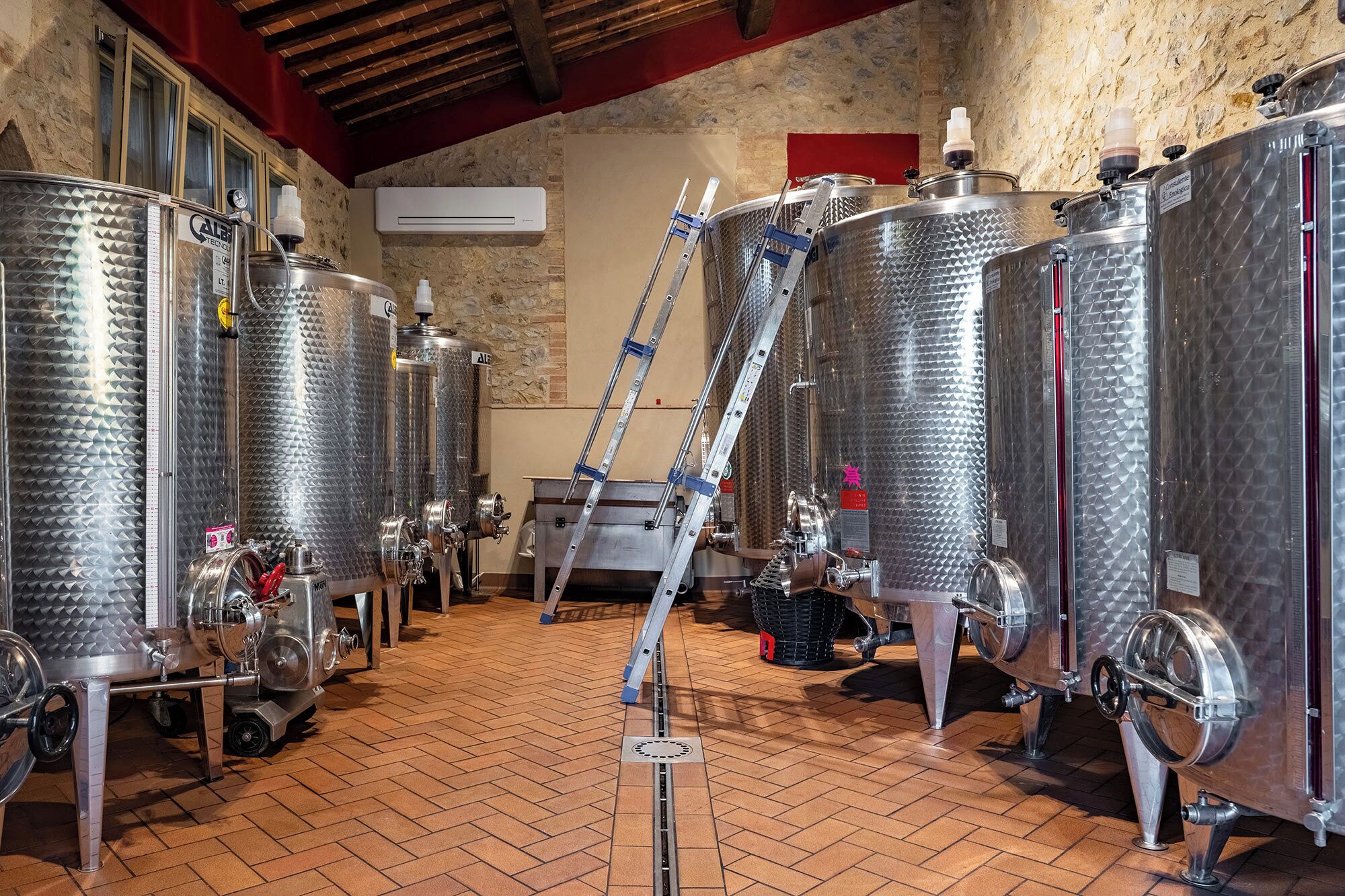 A brewery interior with stainless steel fermentation tanks, a ladder, and a brick floor.