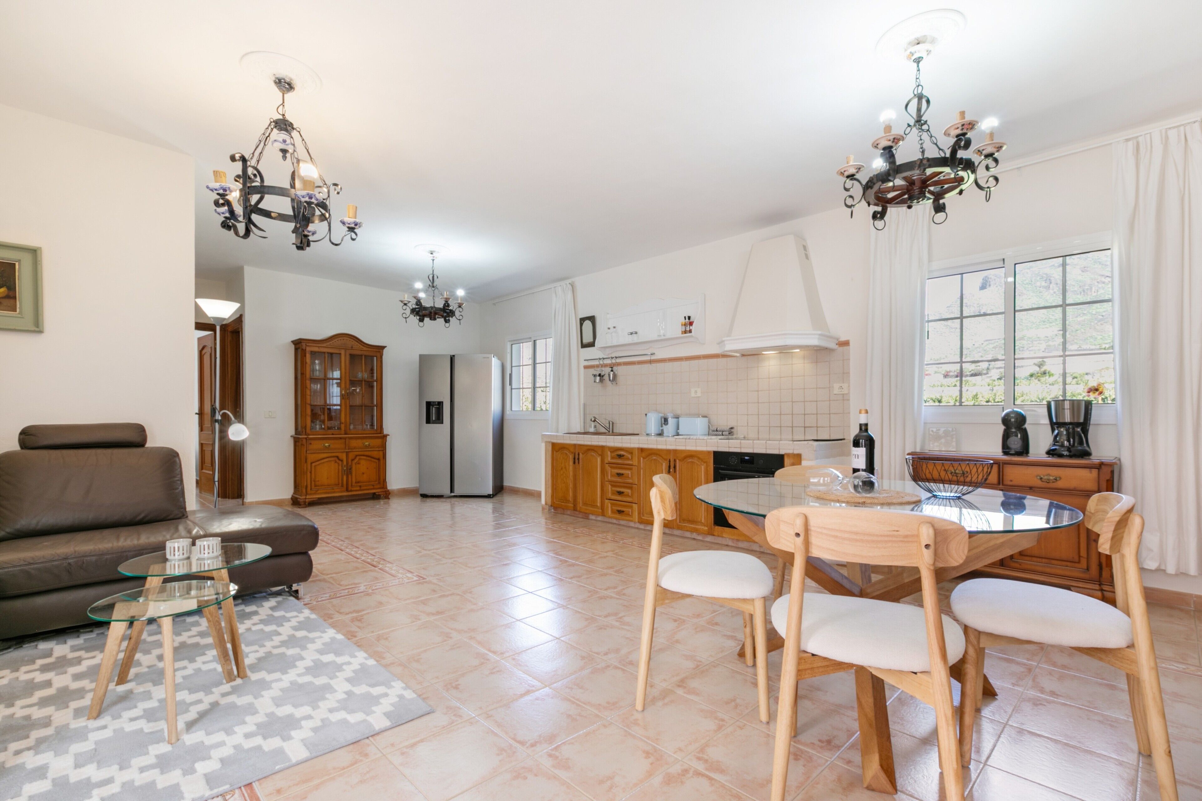 A spacious living area with a sofa, a glass coffee table, and a dining table with chairs. There are two chandeliers, a refrigerator, a kitchen with cabinets, and a window with curtains.