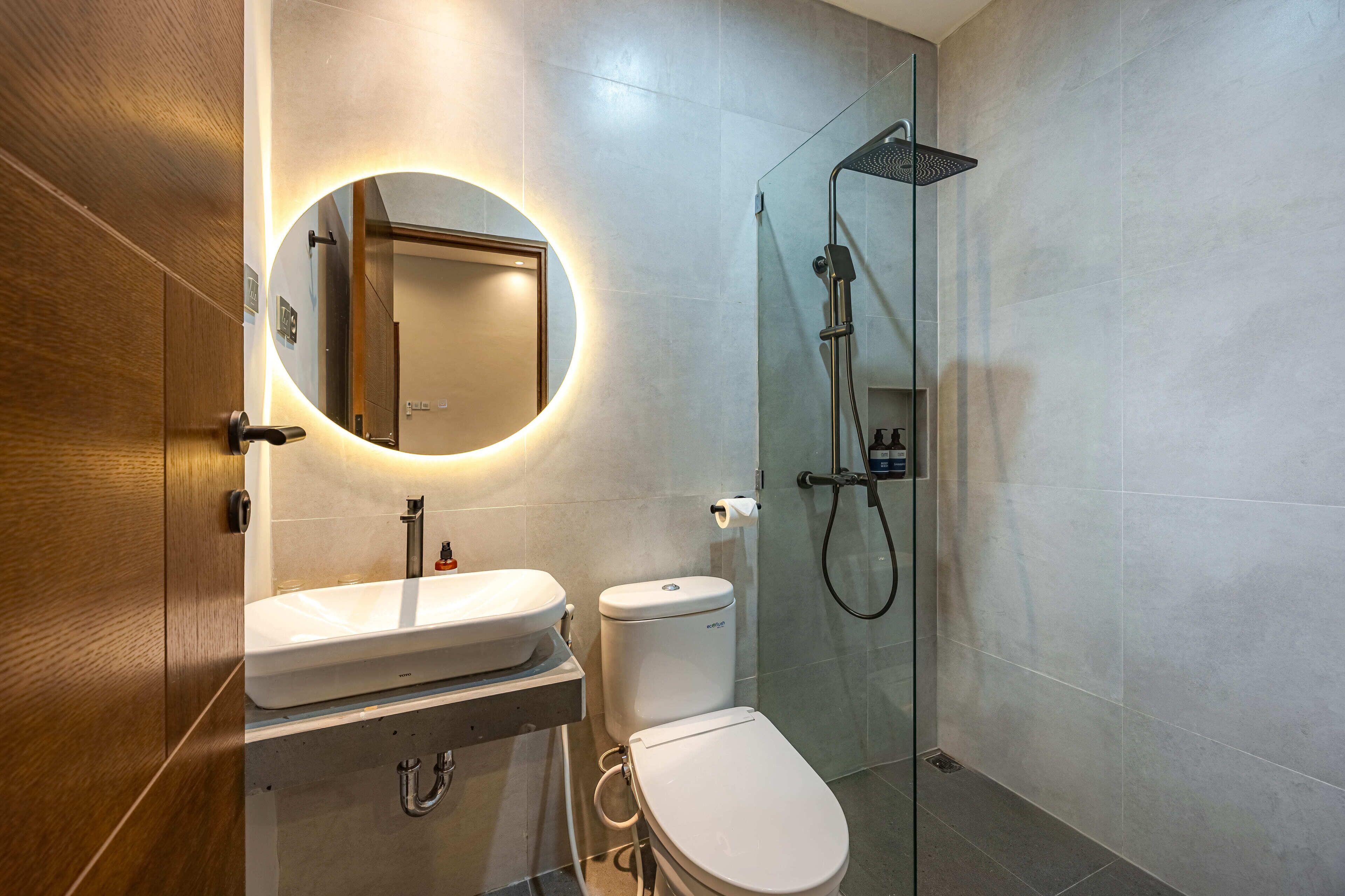 Un baño moderno con puerta de madera, espejo redondo, lavamanos blanco, inodoro y ducha con cerramiento de vidrio.