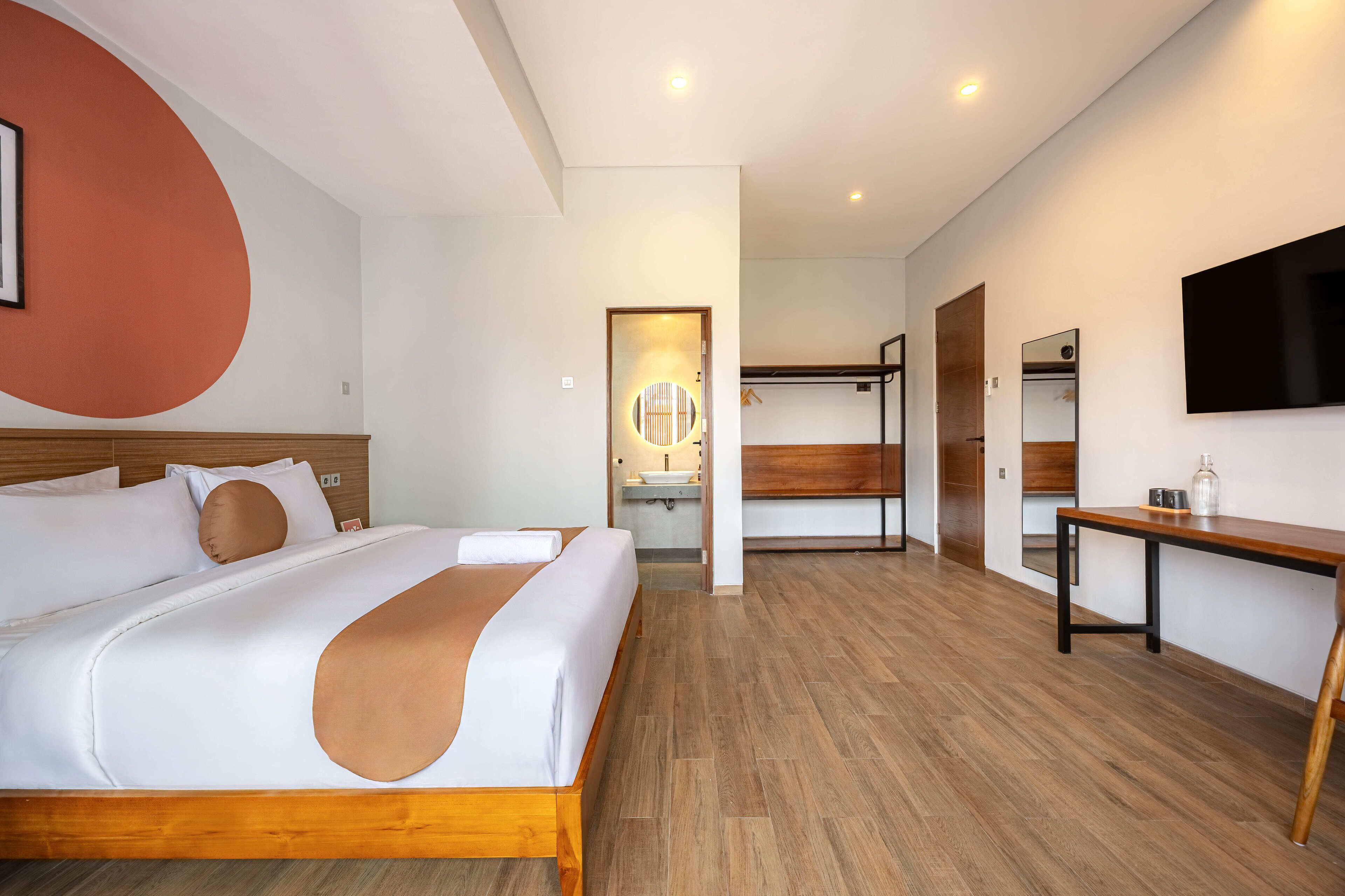 A modern hotel room with a large bed, a wooden headboard, a bedside table, a TV, and a bathroom visible through an open door.