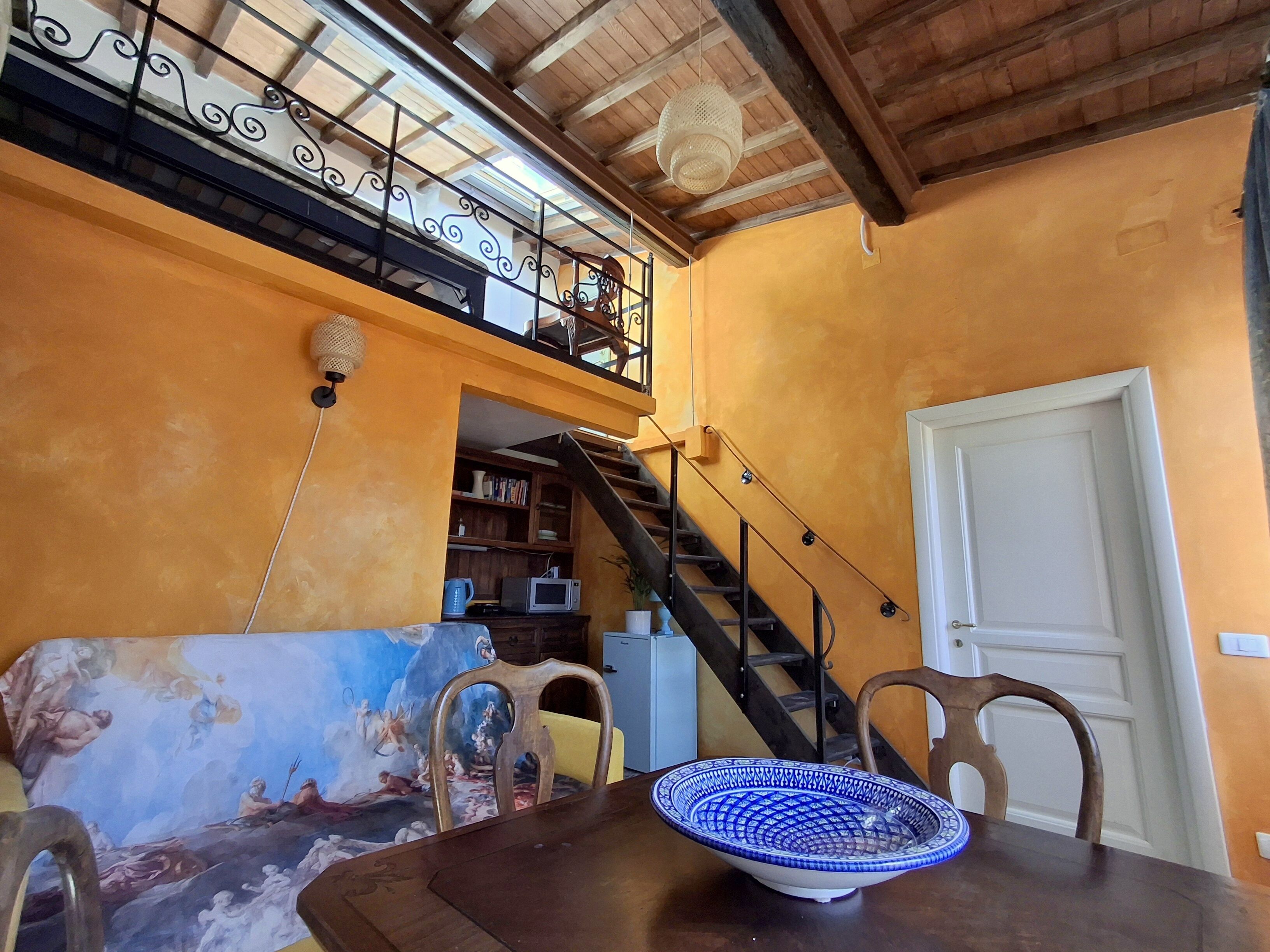 Ein Essbereich mit einem Holztisch, Stühlen und einer blau-weißen, gemusterten Schüssel. Eine Treppe führt zu einer oberen Ebene mit einem Balkon.
