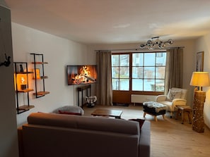 Ein gemütliches Wohnzimmer mit Kamin, einer Couch, einem Sessel, einem Fernseher und einem Fenster mit Vorhängen.