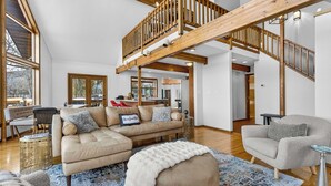 A spacious living area with a high ceiling, wooden beams, a large sofa, a round ottoman, and a staircase leading to an upper level.