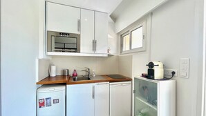 Una cocina compacta con muebles blancos, un lavamanos de acero inoxidable, un refrigerador pequeño y una cafetera.