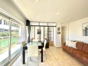 A modern living room with a glass dining table, a brown sofa, and a large window offering a view of a balcony. - copacabana studio (Cannes)