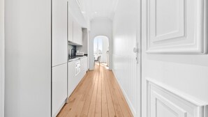 Ein schmaler Korridor mit Holzboden, weißen Wänden und einer Tür zu einem hellen Raum.