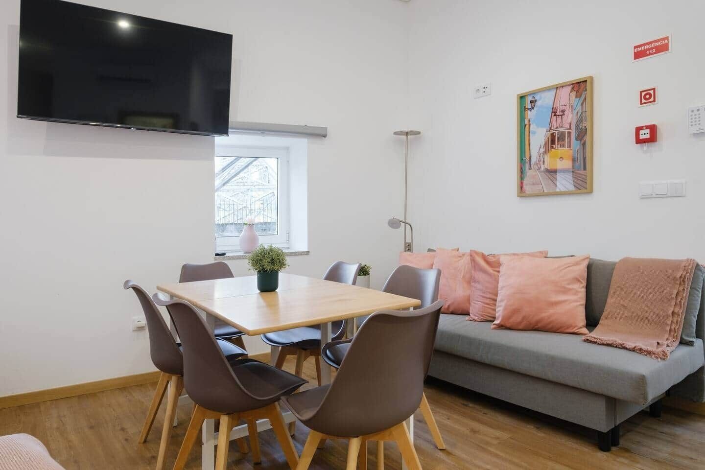 Un saló modern amb zona de menjador, un sofà gris amb coixins rosa, un televisor de pantalla plana fixat a la paret i una finestra amb vista a un edifici.