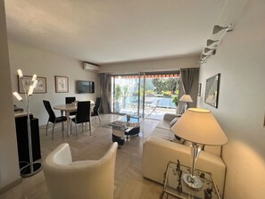 A modern living room with a sofa, coffee table, and dining area. There is a TV mounted on the wall and a balcony with plants. - Alexandra appartement (Cannes)